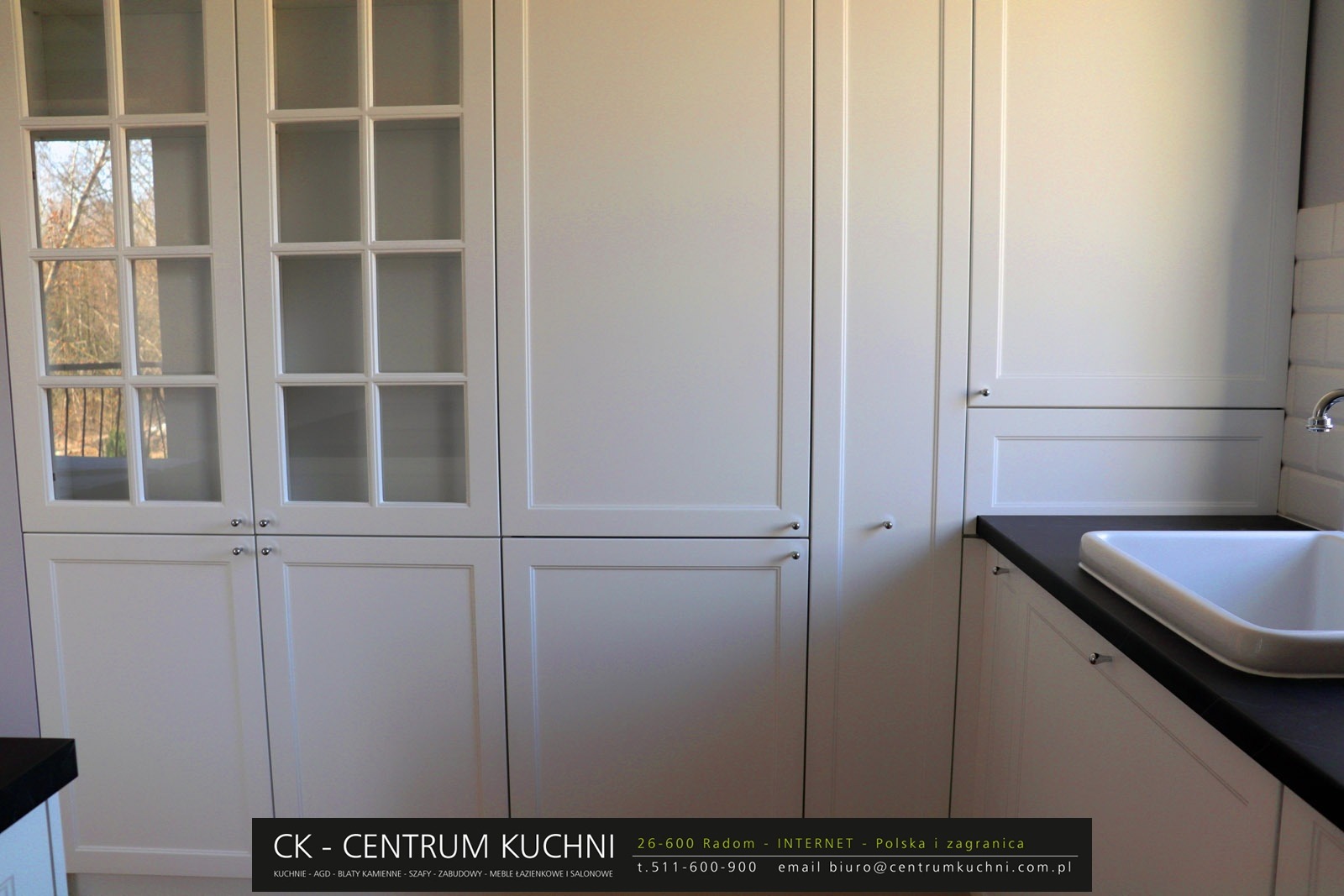 Biała zabudowa kuchenna z frezowanymi frontami, przeszklone szafki górne z widokiem na drzewa za oknem, czarny blat i biały zlew, minimalistyczna bateria.