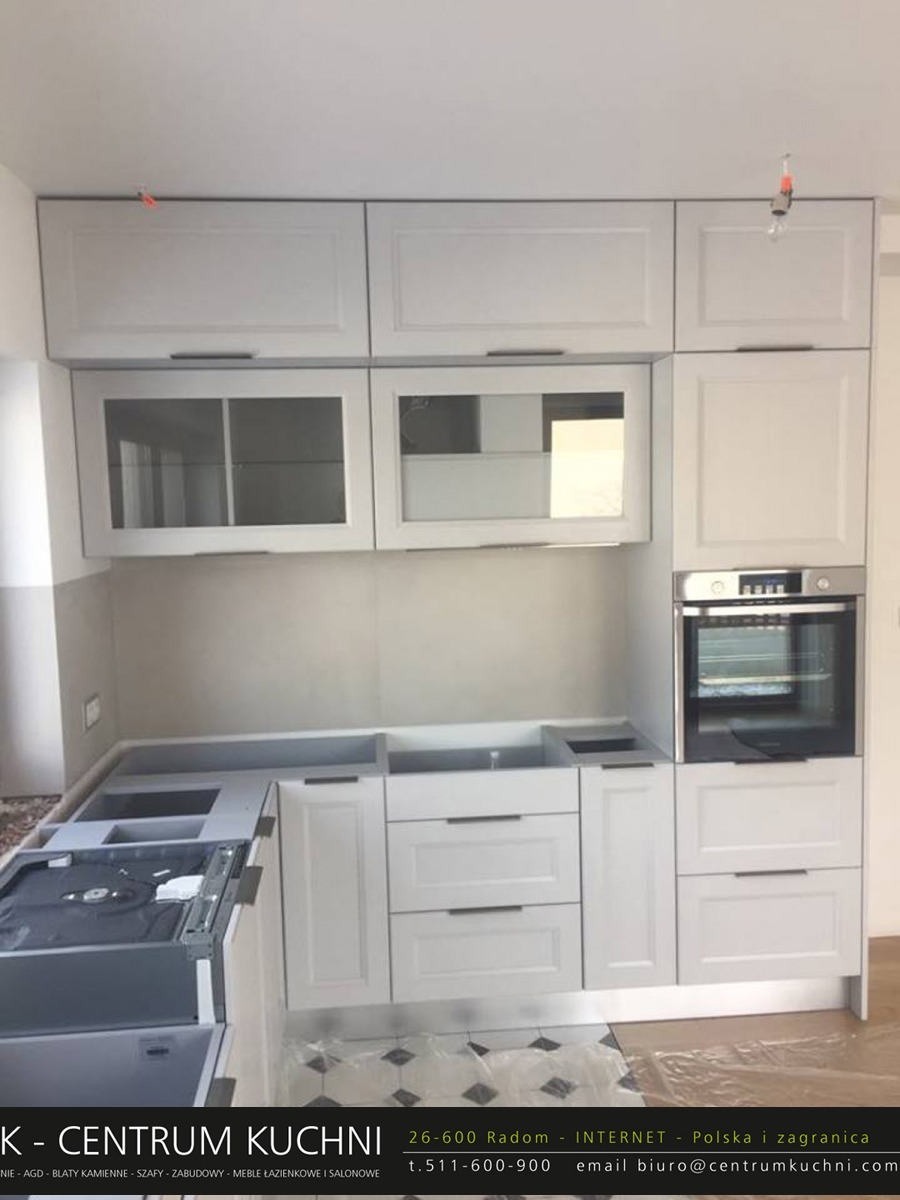 Wnętrze kuchni w trakcie montażu, widoczne białe szafki z szufladami i przeszklonymi górnymi szafkami, brak blatu, widoczny piekarnik, podłoga zabezpieczona folią.
