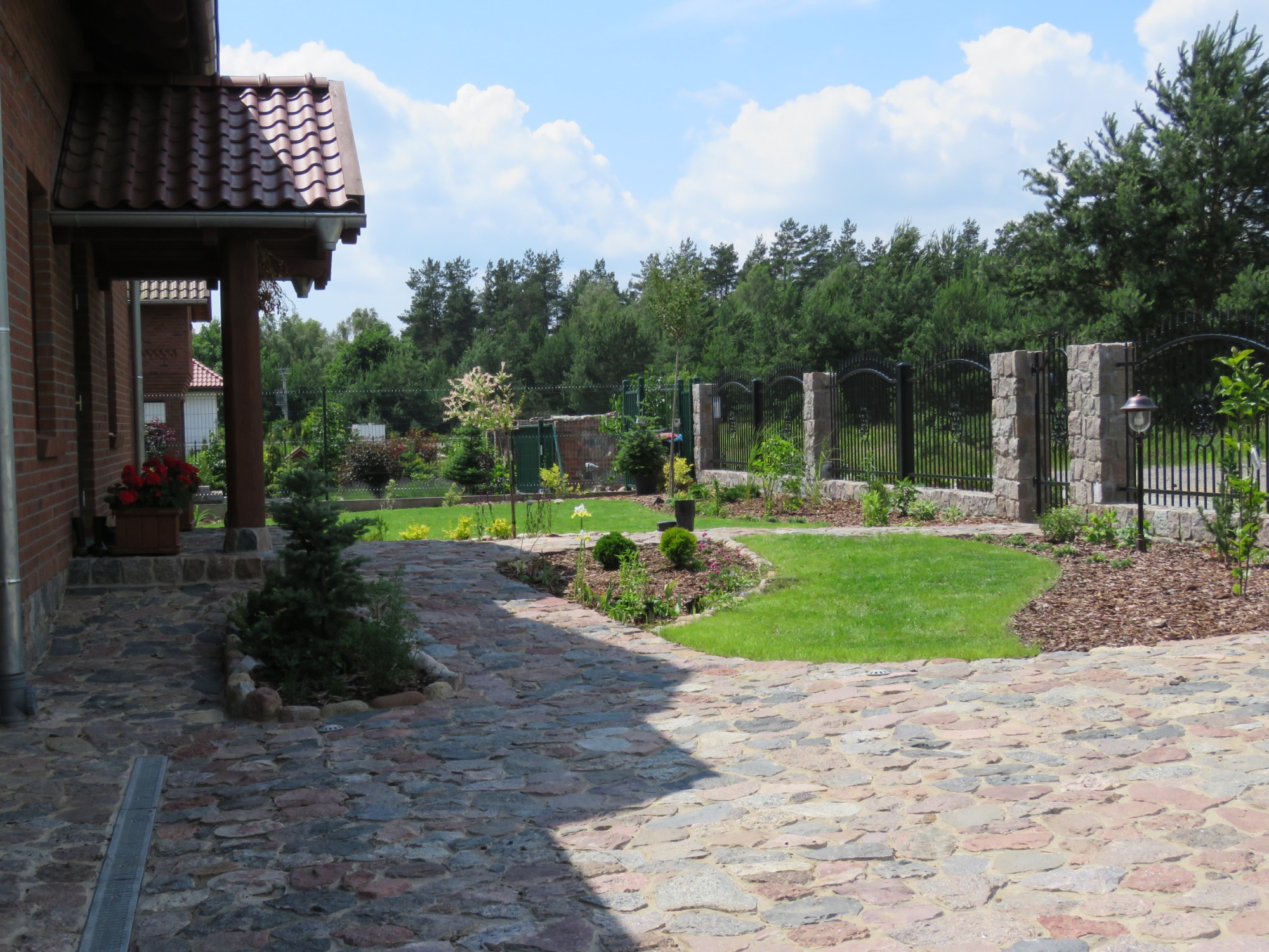 Kamienna ścieżka prowadząca przez zadbany ogród z równo przystrzyżonym trawnikiem, niskimi krzewami i drzewami w tle, ogrodzony kutym płotem z kamiennymi słupkami.