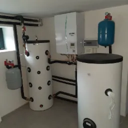 Zestaw urządzeń w kotłowni: biała jednostka zewnętrzna z zielonym akcentem marki Ecodan Mitsubishi Electric, dwa białe zbiorniki akumulacyjne Kospel z czarnymi okrągłymi otworami rewizyjnymi...