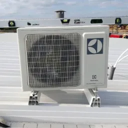 Biała jednostka zewnętrzna klimatyzacji Electrolux na dachu, z poziomicą Stanley FatMax na górze, widoczne kable i elementy montażowe. Montaż w toku.