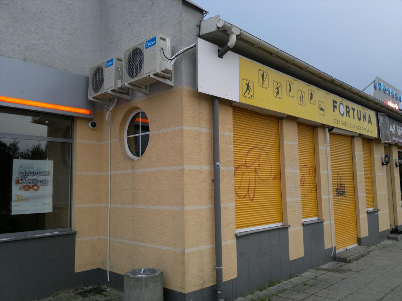 Dwa klimatyzatory Midea zamontowane na elewacji budynku Fortuna zakłady bukmacherskie z żółtymi roletami. Widoczny fragment okna i okrągłe okno.