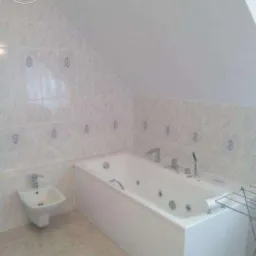 Biała wanna z hydromasażem i bidet w łazience z ukośnym sufitem i jasnymi, wzorzystymi płytkami.