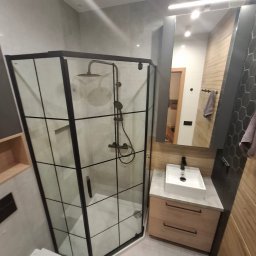 Dominik Filak - Nowoczesna łazienka z kabiną prysznicową typu walk-in z czarnymi profilami, umywalką nablatową i drewnianymi akcentami. Widoczne szare płytki i oświetlenie LED.