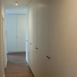 Długi korytarz z rzędem białych szaf z czarnymi gałkami, widoczne drewniane panele podłogowe i szafa w głębi.