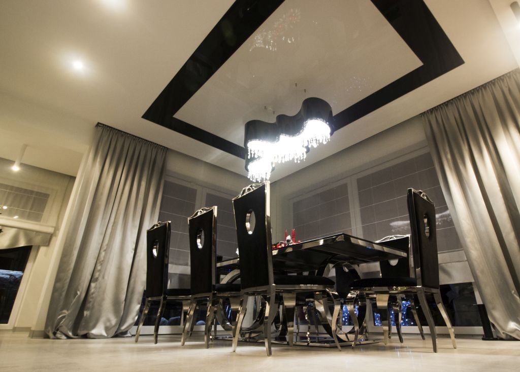 Elegancka jadalnia z czarnym stołem i krzesłami, błyszczący sufit z czarną obwódką i nowoczesnym żyrandolem, okna zasłonięte srebrnymi zasłonami.