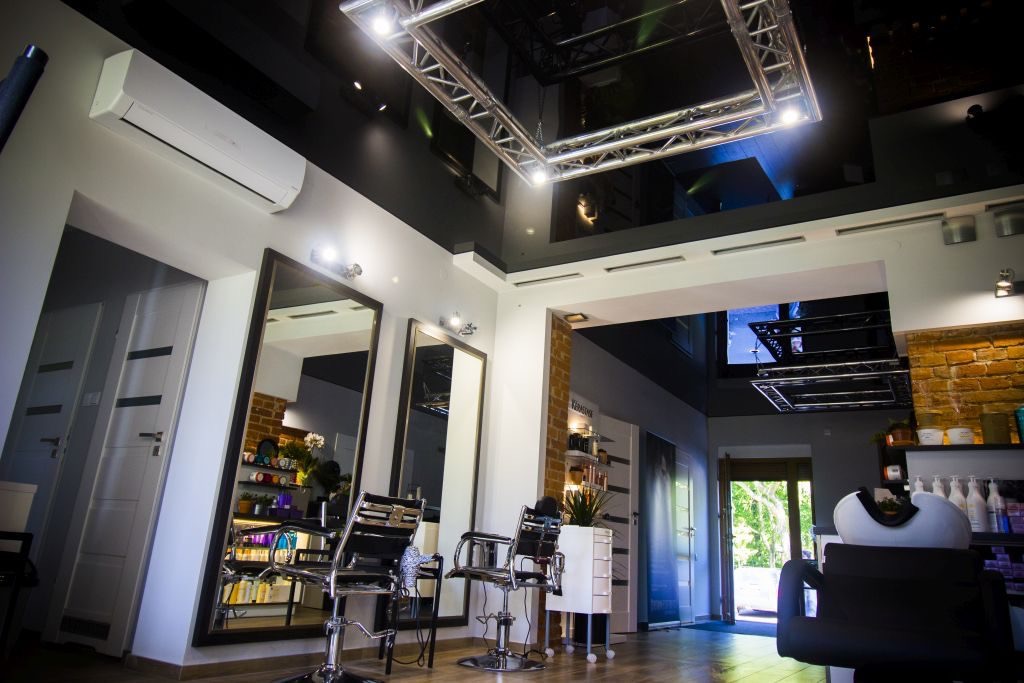 Nowoczesny salon fryzjerski z czarnym sufitem napinanym, podświetlonym metalową konstrukcją oświetleniową, widoczne lustra i krzesła fryzjerskie.