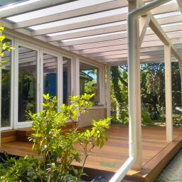 Tarasy z montażem i drewno do Sauny - Zadaszony taras z egzotycznego drewna Angelim Amorgosso, z białymi ramami i przeszklonymi drzwiami, otoczony zielenią ogrodu.