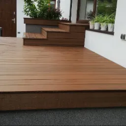 Nowoczesny taras z deski kompozytowej w kolorze brązowym, z niskimi schodami prowadzącymi do drzwi, donice z roślinami przy oknie, widok z bliska na fakturę deski.