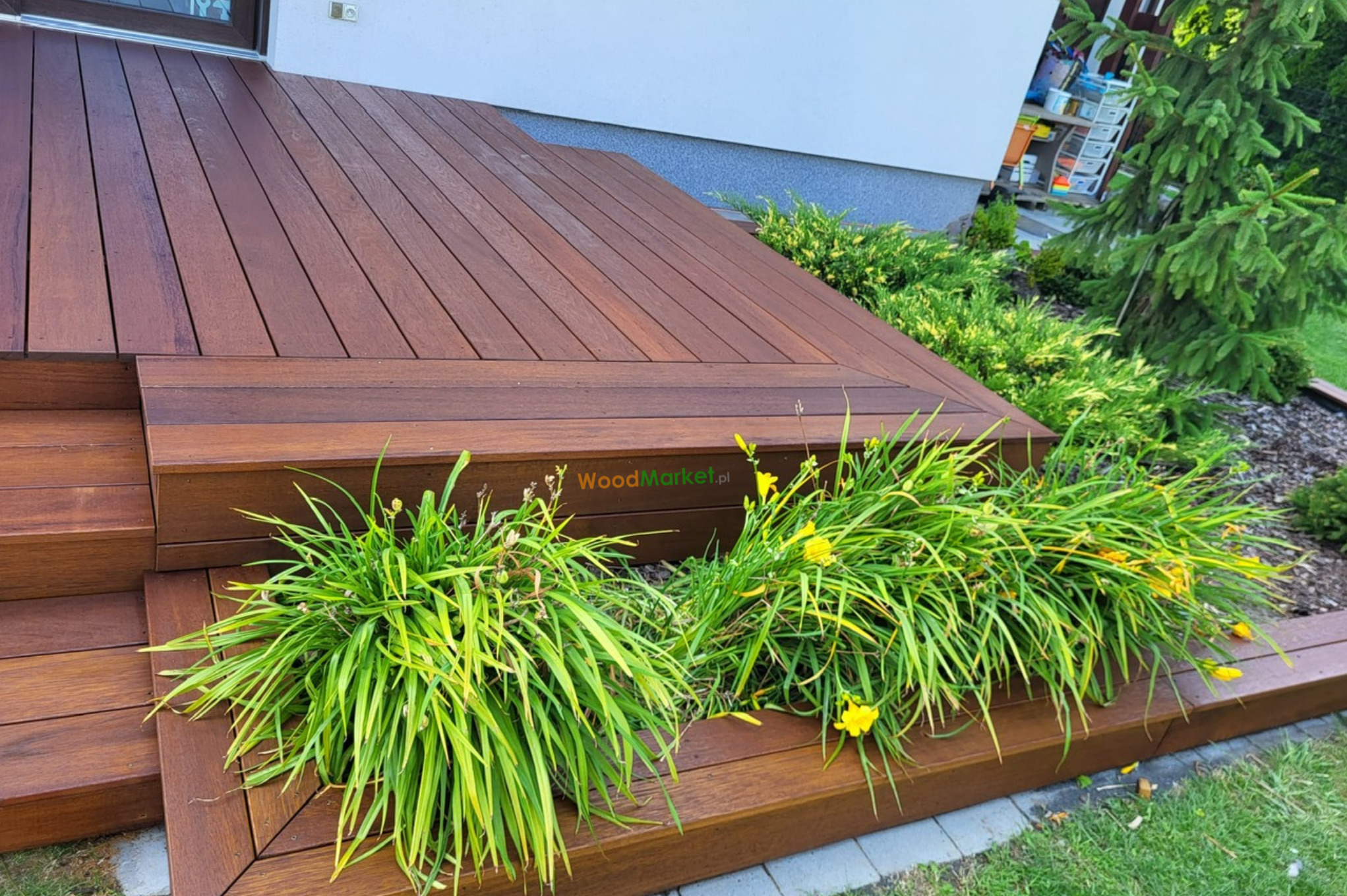 Ciemnobrązowy taras z desek, z niskimi schodami i rabatą z żółtymi liliowcami, przylegający do jasnego budynku.