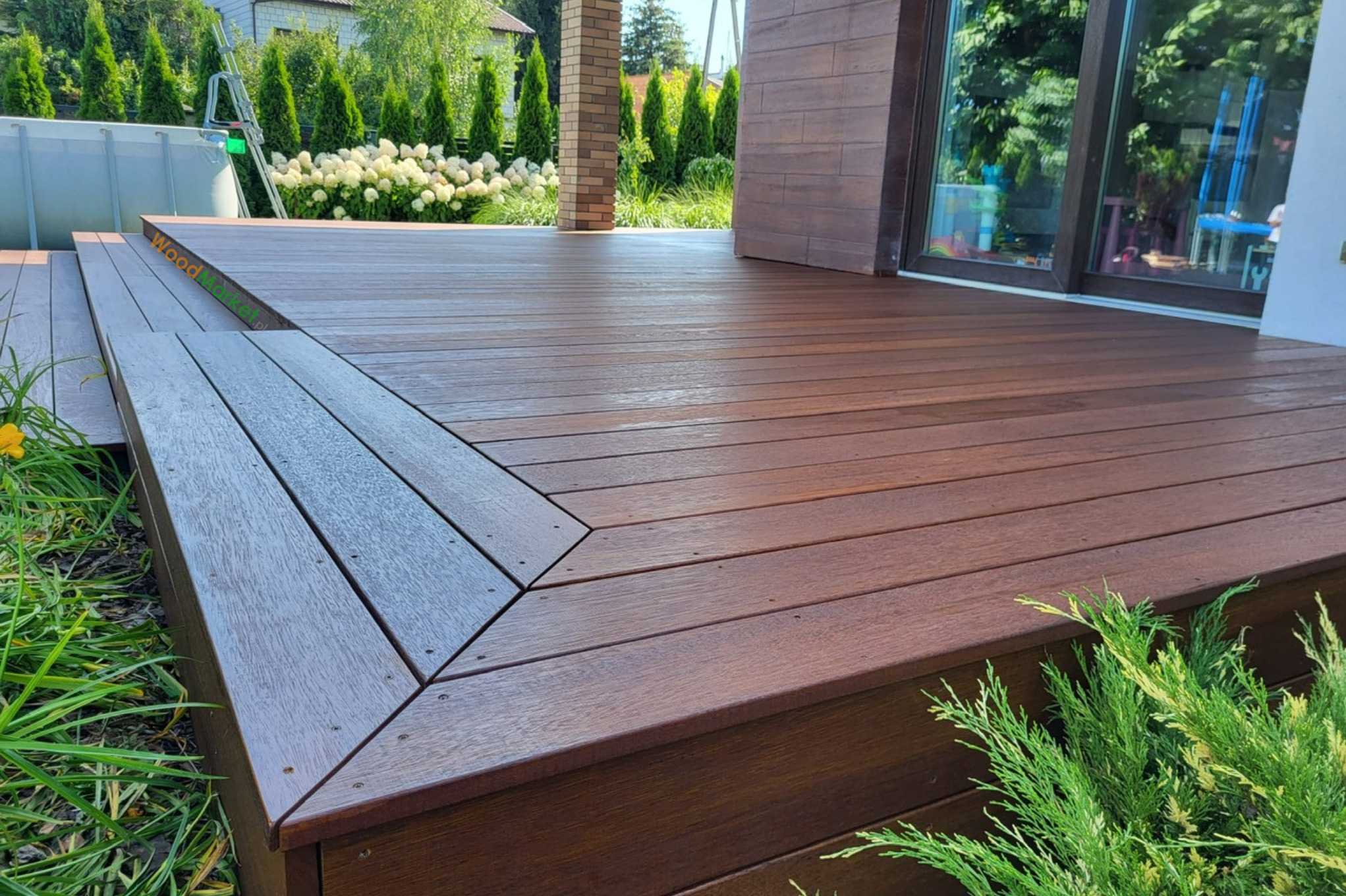 Nowoczesny taras z ciemnego drewna z widocznym przejściem na wyższy poziom, otoczony zielenią i kwiatami, z basenem w tle.