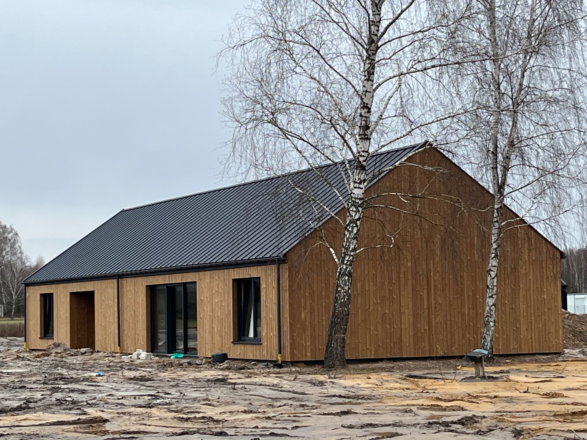 Nowoczesny dom z elewacją z pionowych desek, czarnym dachem i dużymi oknami, otoczony błotnistym terenem w trakcie prac wykończeniowych.