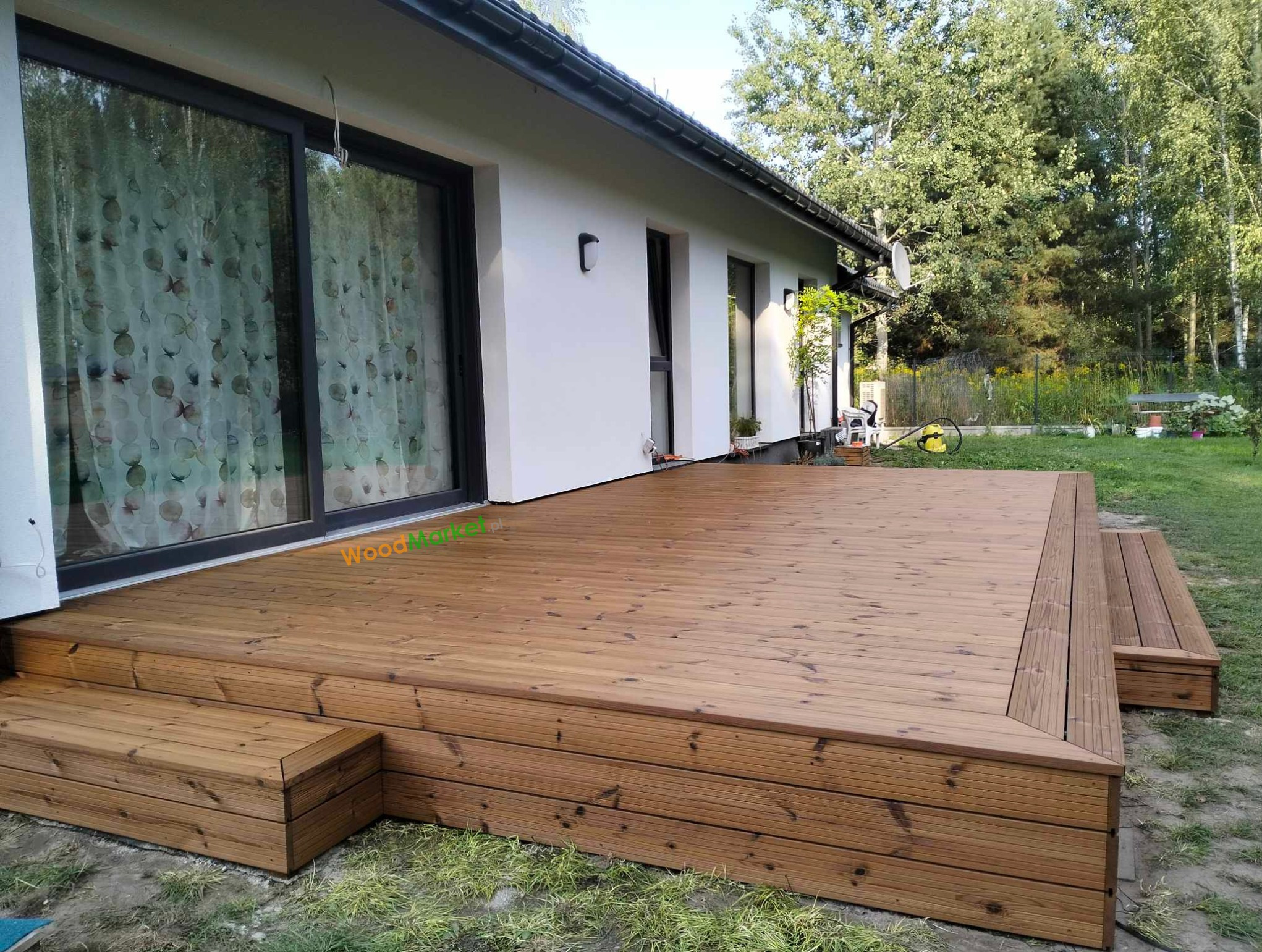 Nowoczesny taras z drewna z niskimi stopniami, przylegający do białego domu z dużymi przesuwnymi drzwiami tarasowymi.