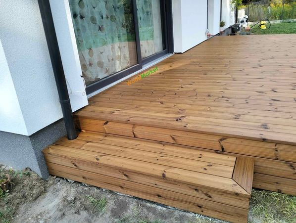 Drewniany taras z dwoma stopniami, przylegający do białej ściany domu z dużymi przesuwnymi drzwiami, widoczny fragment rynny.