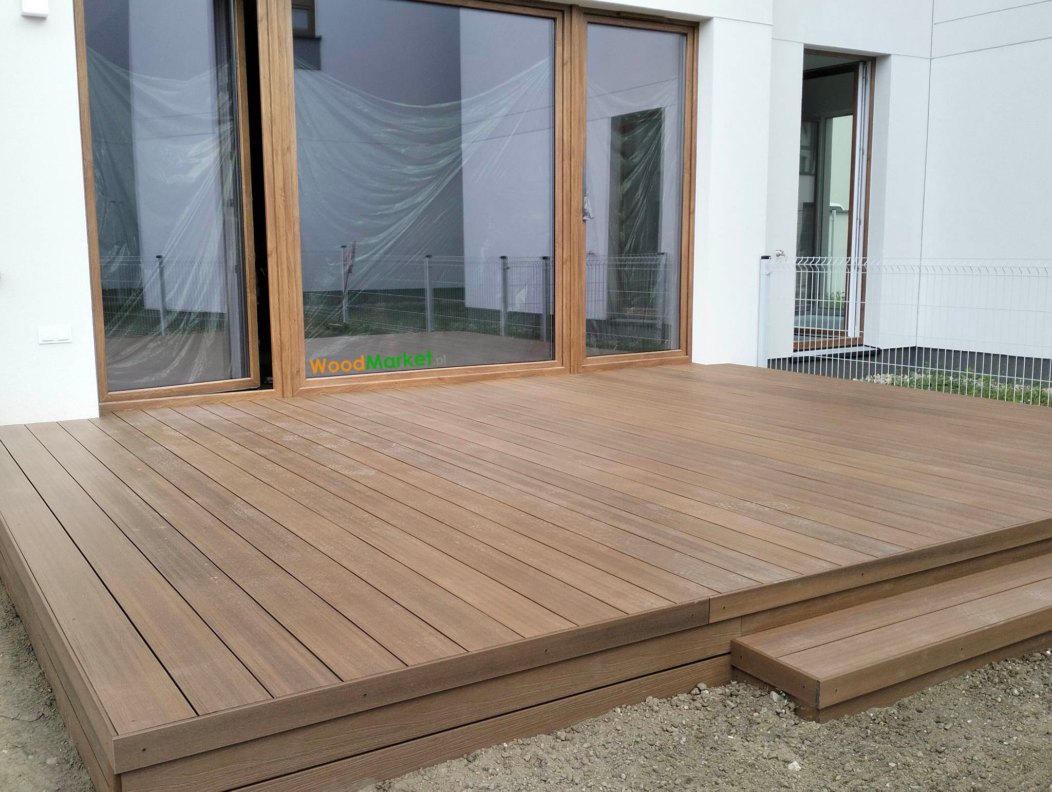 Nowoczesny taras kompozytowy w odcieniach brązu, z dwoma stopniami, przylegający do budynku z dużymi przesuwnymi oknami, częściowo zasłoniętymi folią ochronną.