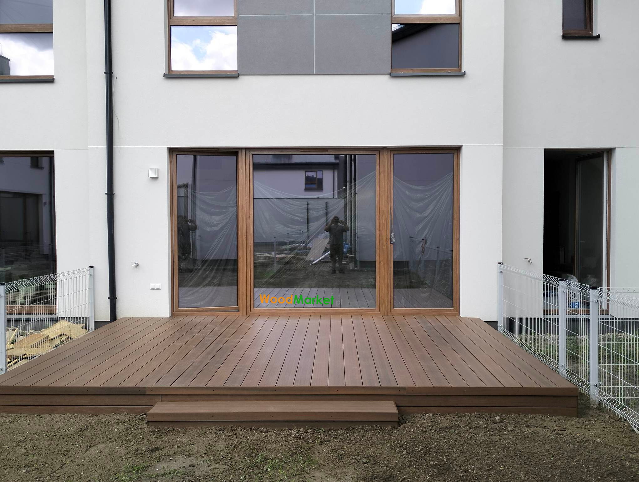 Nowoczesny taras z desek kompozytowych w kolorze brązowym, z dwoma stopniami, przylegający do białego budynku z dużymi oknami, częściowo zasłoniętymi folią ochronną.