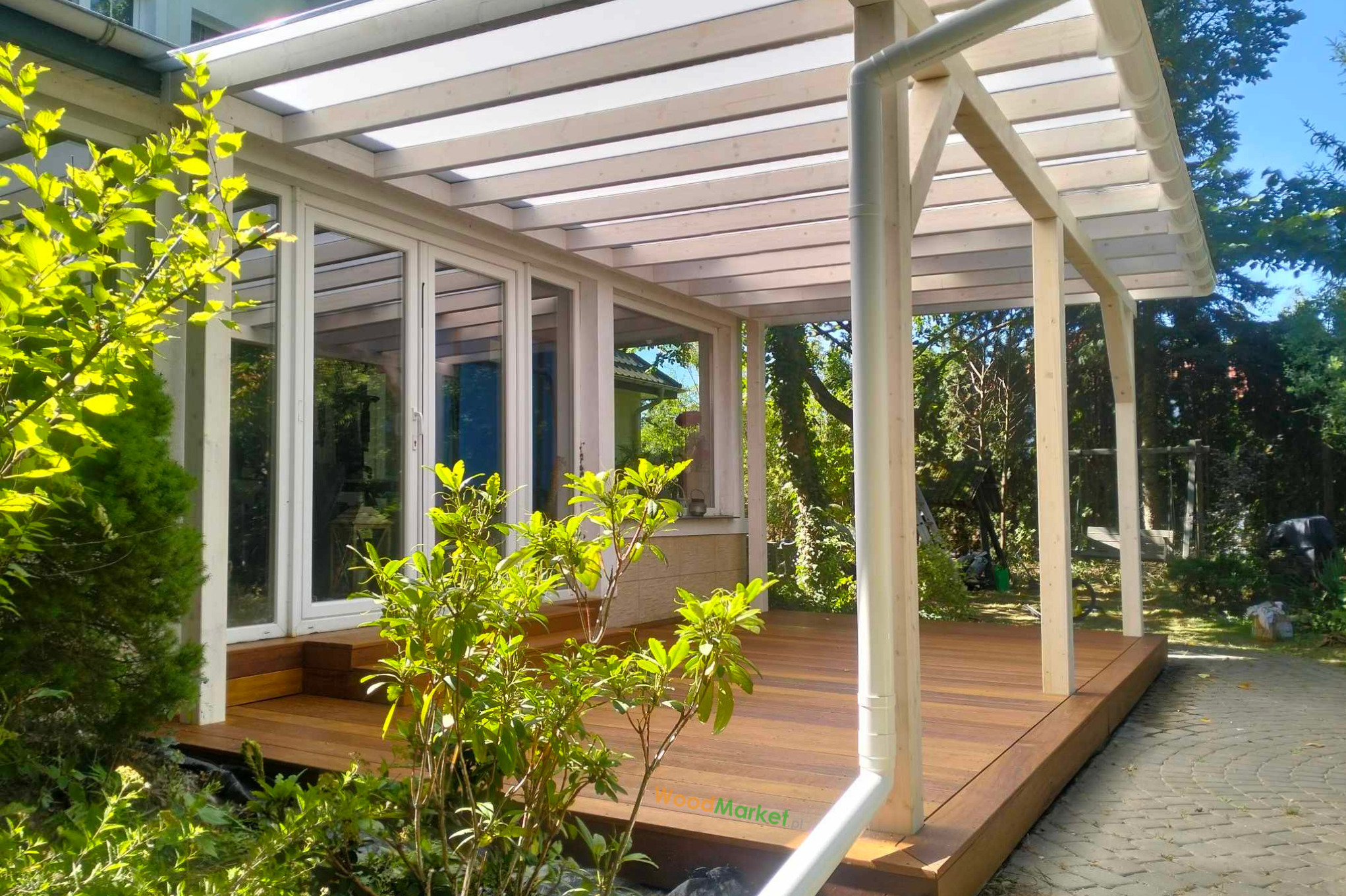 Zadaszony taras z egzotycznego drewna Angelim Amorgosso, z białymi ramami i przeszklonymi drzwiami, otoczony zielenią ogrodu.