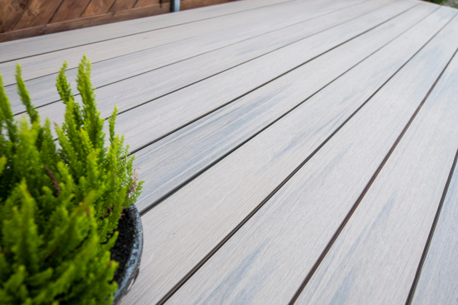 Jasnoszara deska kompozytowa Gardin ułożona na tarasie, z doniczką z zieloną rośliną w lewym dolnym rogu, widok z bliska pod kątem.