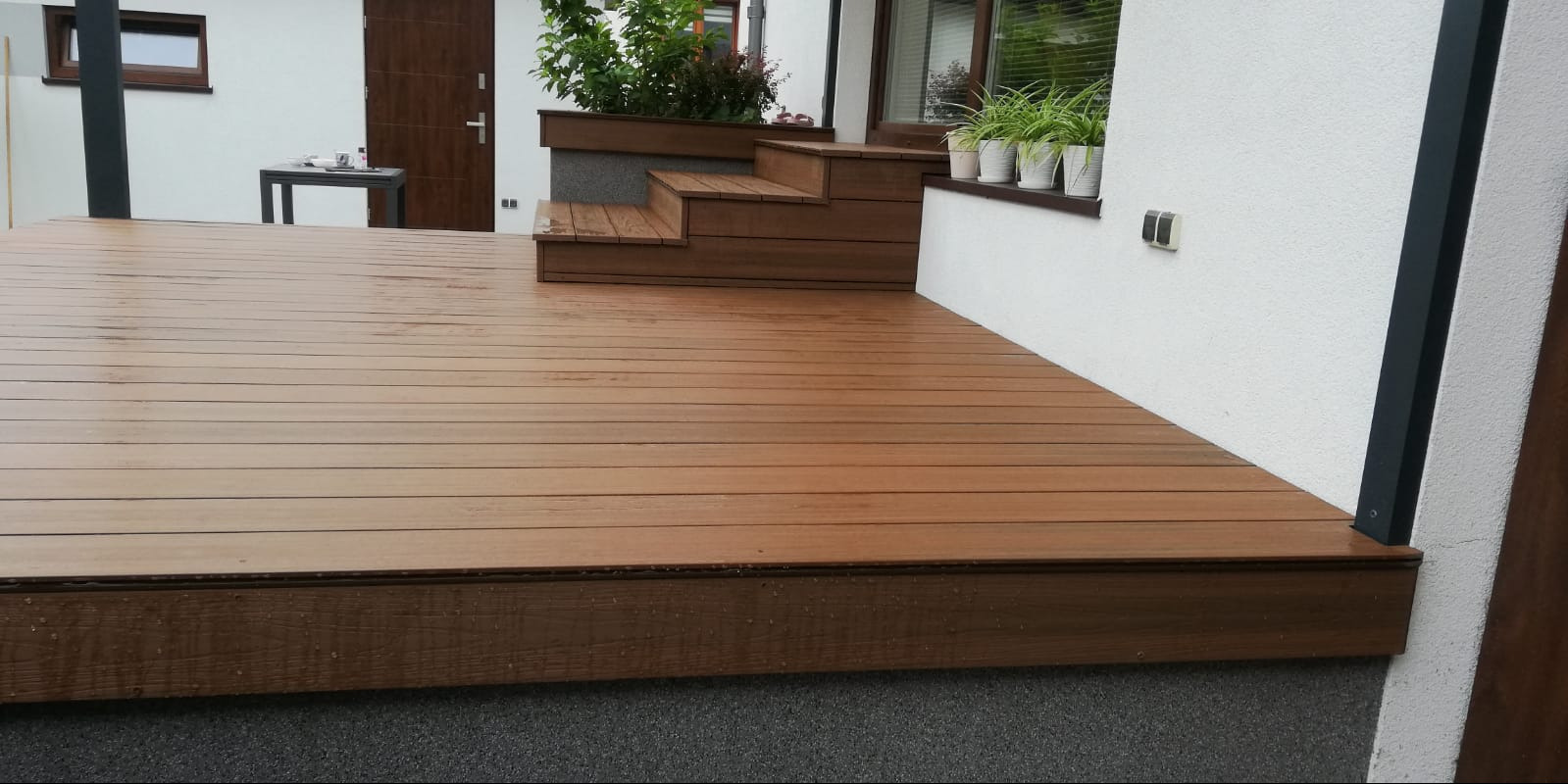 Nowoczesny taras z deski kompozytowej w kolorze brązowym, z niskimi schodami prowadzącymi do drzwi, donice z roślinami przy oknie, widok z bliska na fakturę deski.