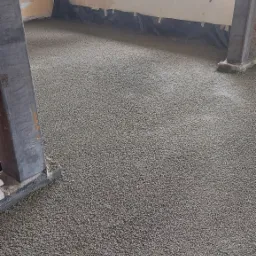 Świeżo wylany styrobeton na podłodze w pomieszczeniu z widocznym oknem i czarną folią przy ścianach, przygotowany pod dalsze prace wykończeniowe.