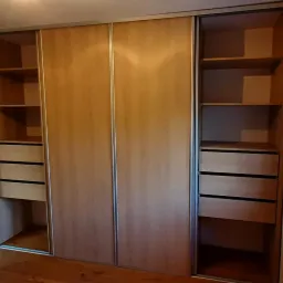 Jasna, drewnopodobna szafa przesuwna z otwartymi bokami, ukazującymi półki i szuflady, z metalowymi prowadnicami, w jasnym wnętrzu.