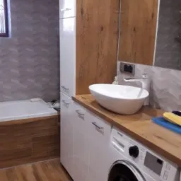 Białe szafki łazienkowe z drewnianym blatem, na którym stoi owalna umywalka nablatowa i pralka automatyczna. W tle widoczna wanna obudowana drewnem oraz wysoka szafka z drewnianymi frontami.