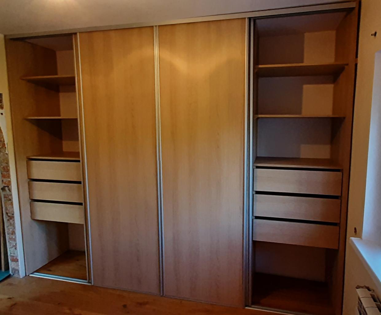 Jasna, drewnopodobna szafa przesuwna z otwartymi bokami, ukazującymi półki i szuflady, z metalowymi prowadnicami, w jasnym wnętrzu.