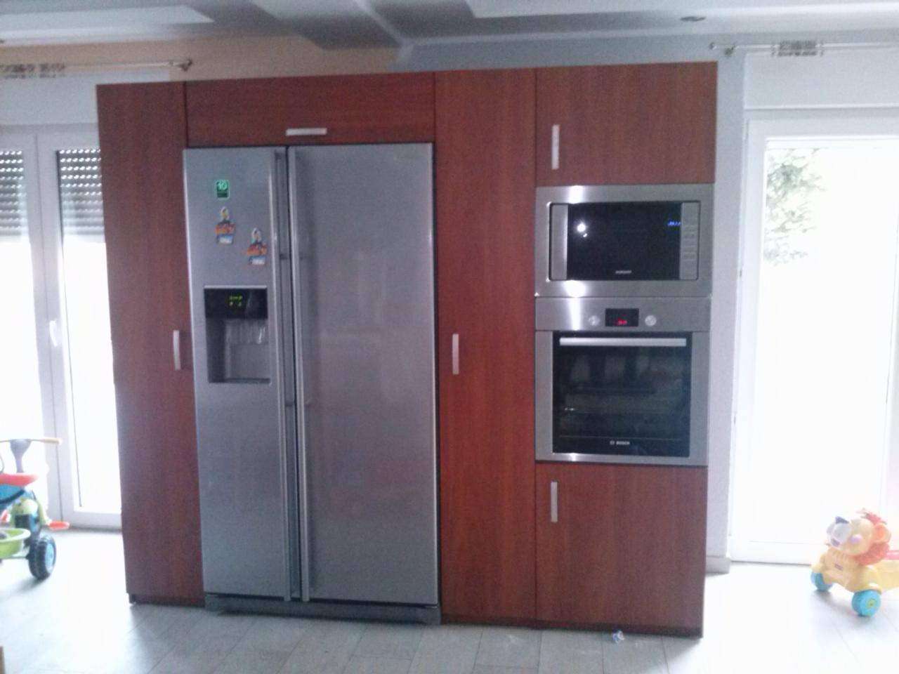 Zabudowa kuchenna w odcieniach brązu z wbudowanym sprzętem AGD: lodówką z kostkarką, piekarnikiem i mikrofalówką; na podłodze zabawki dziecięce.