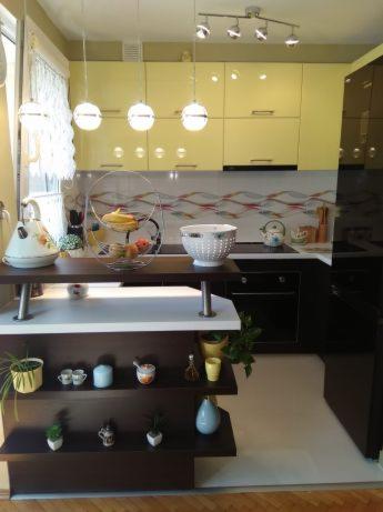 Żółta kuchnia z wyspą i trzema wiszącymi lampami kulami, ozdobiona półkami z dekoracjami i owocami