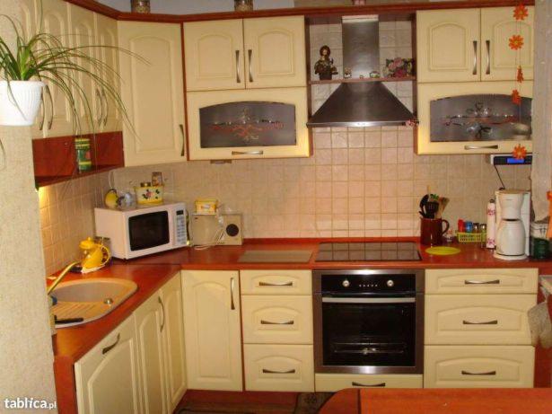 Jasna kuchnia z kremowymi szafkami i czerwonymi blatami, wyposażona w kuchenkę elektryczną, piekarnik, mikrofalówkę i ekspres do kawy, z dekoracyjnymi elementami na górnych szafkach.