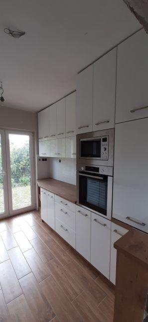 Nowoczesna kuchnia z białymi, lakierowanymi frontami, drewnianym blatem i wbudowanym piekarnikiem oraz mikrofalówką, widok z perspektywy podłogi z płytek imitujących drewno, widoczne niedokończone...