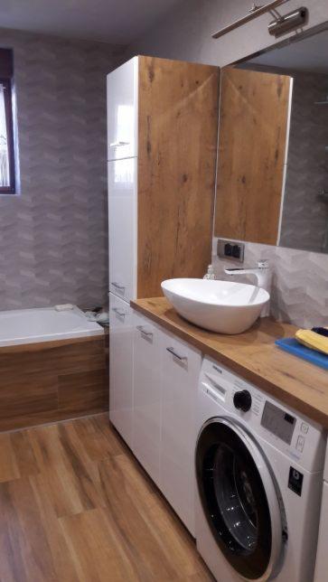 Białe szafki łazienkowe z drewnianym blatem, na którym stoi owalna umywalka nablatowa i pralka automatyczna. W tle widoczna wanna obudowana drewnem oraz wysoka szafka z drewnianymi frontami.