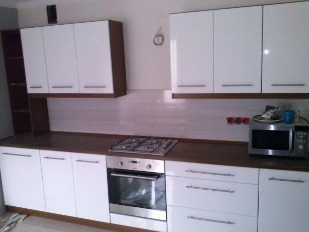 Nowoczesna kuchnia z białymi frontami szafek i ciemnobrązowym blatem, widoczna płyta gazowa i piekarnik w zabudowie, nad blatem wiszące szafki z białymi frontami i ciemnobrązowymi korpusami, po...