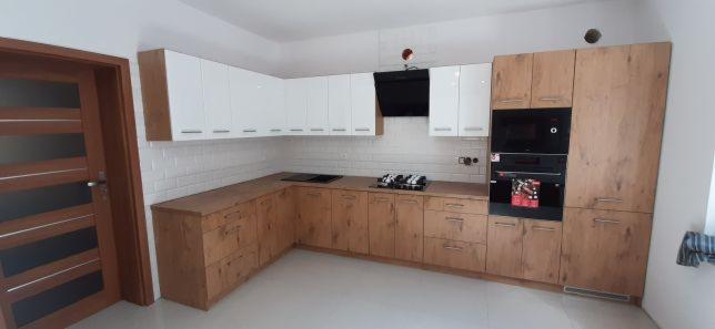 Nowoczesna kuchnia na wymiar z drewnianymi frontami dolnymi i białymi górnymi szafkami, czarny okap, białe kafelki na ścianie i brązowe drzwi z przeszkleniami.