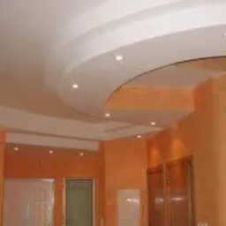 Kremowy sufit podwieszany z dekoracyjnymi łukami i punktowym oświetleniem w pomarańczowym wnętrzu. Widoczne fragmenty ścian z fakturą i drzwiami.