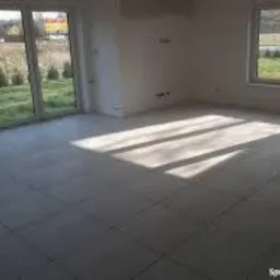 Jasne, nowo wykafelkowane pomieszczenie z dużymi oknami i widokiem na zewnątrz. Światło słoneczne tworzy geometryczne wzory na podłodze. Widoczne niedoskonałości na ścianach wskazują na trwający...