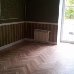 Jasny parkiet ułożony w jodełkę w pokoju z oknem wychodzącym na ogród. Na ścianach dekoracyjne panele i grzejnik. Widoczne światło dzienne.