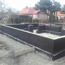 Świeżo zaizolowane, pomalowane na czarno fundamenty domu jednorodzinnego w trakcie budowy, z widocznymi prętami zbrojeniowymi i fragmentami szalunków w wykopie.