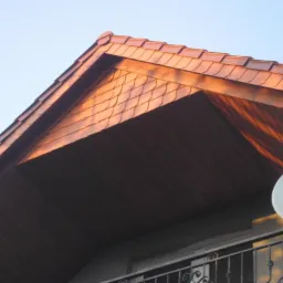 Fragment dachu z brązową dachówką karpiówką, widoczny z dołu, z balkonem i anteną satelitarną w kadrze. Oświetlenie popołudniowe.