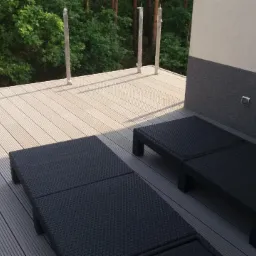 Beżowy taras z desek kompozytowych z dwoma czarnymi leżakami, słupki z folią ochronną, widok na zieleń drzew