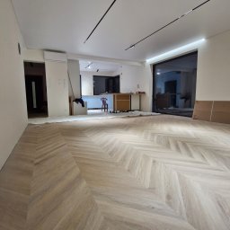 Usługi Remontowo Budowlane '' WalBud '' Walura Łukasz - Jasna, drewniana podłoga ułożona we wzór jodełki francuskiej w przestronnym, częściowo umeblowanym wnętrzu. Widoczne oświetlenie szynowe i duże okno.
