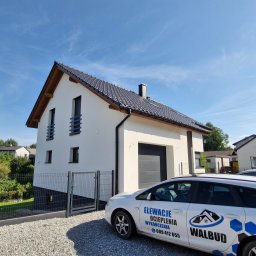 Usługi Remontowo Budowlane '' WalBud '' Walura Łukasz - Biały dom jednorodzinny z szarą dachówką i elementami drewnianymi, widoczny z zewnątrz, z samochodem firmowym z logo na pierwszym planie.