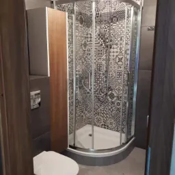 Wykończona łazienka z kabiną prysznicową narożną, obudowaną płytkami w czarno-białe wzory patchworkowe, z toaletą wiszącą i szafką w odcieniach brązu i szarości.