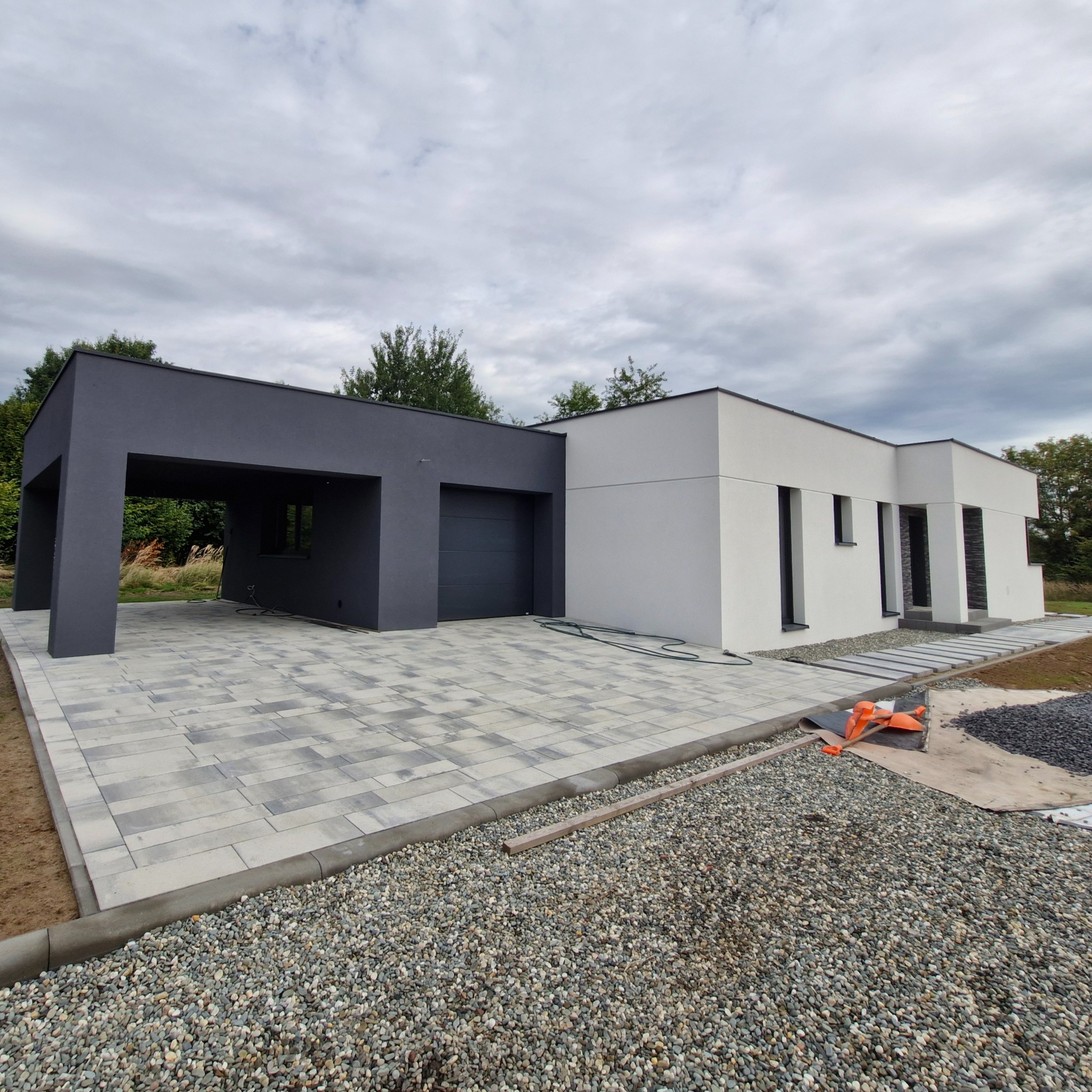 Nowoczesny dom jednorodzinny z szarą elewacją i garażem, wykończony jasnym tynkiem. Ułożona kostka brukowa na podjeździe, widoczne prace wykończeniowe wokół budynku.