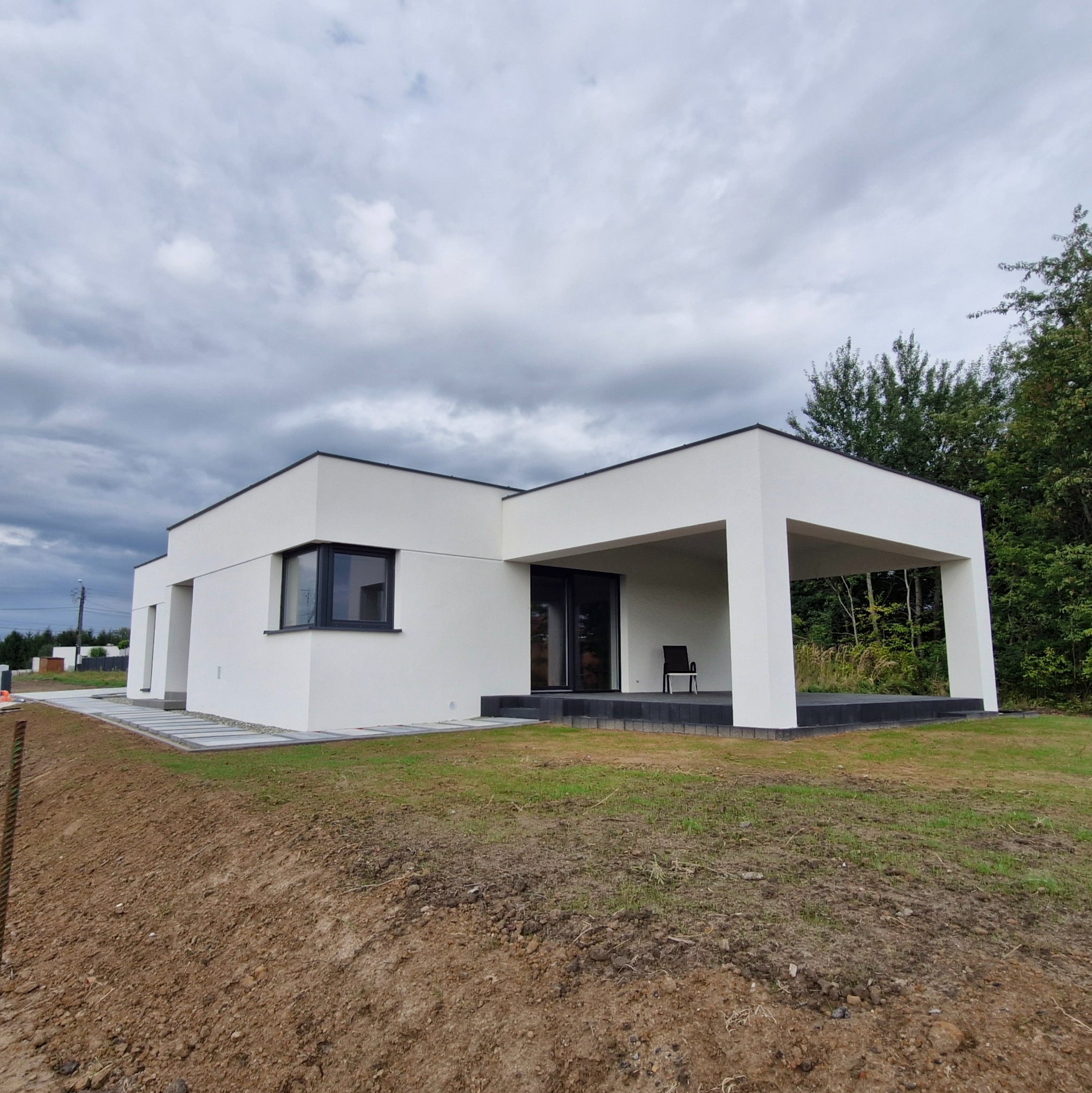 Nowoczesny, parterowy dom z białymi ścianami i płaskim dachem, widoczny z perspektywy ogrodu. Minimalistyczny design, duże okna, ciemna stolarka okienna i drzwiowa.