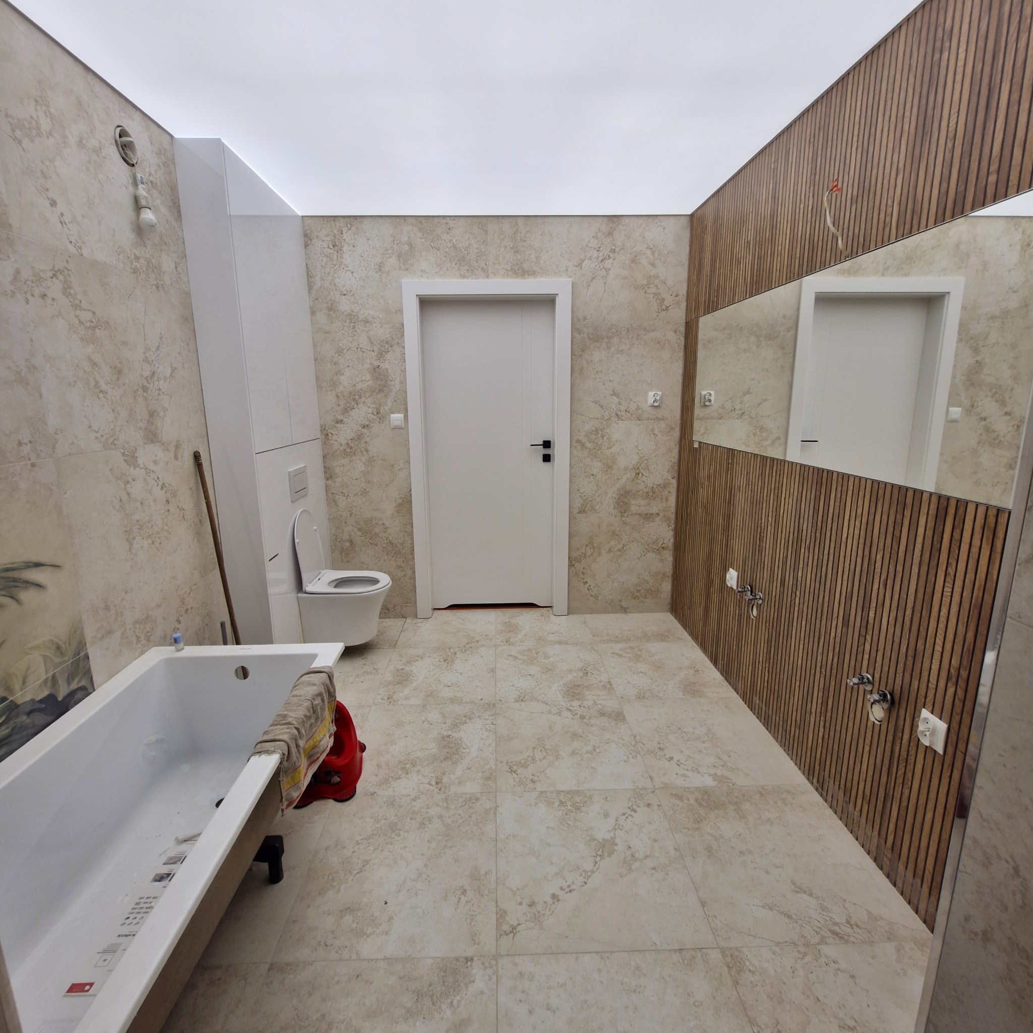 Nowoczesna łazienka w trakcie wykańczania: beżowe płytki, drewniane panele ścienne, biała wanna i toaleta. Widoczne niedokończone instalacje.