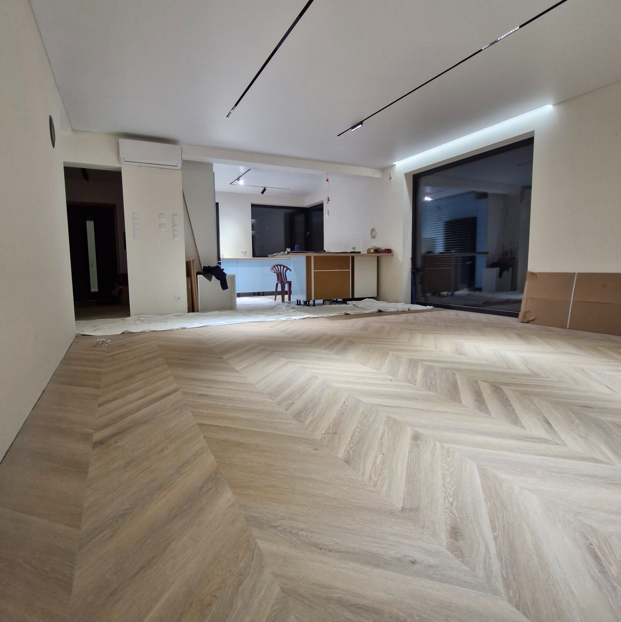 Jasna, drewniana podłoga ułożona we wzór jodełki francuskiej w przestronnym, częściowo umeblowanym wnętrzu. Widoczne oświetlenie szynowe i duże okno.