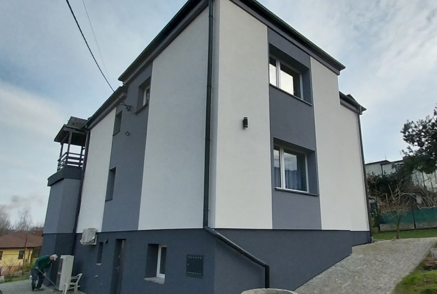 Dwukondygnacyjny dom po renowacji elewacji w odcieniach szarości i bieli, z widocznym fragmentem ogrodu i pracującym mężczyzną w tle.