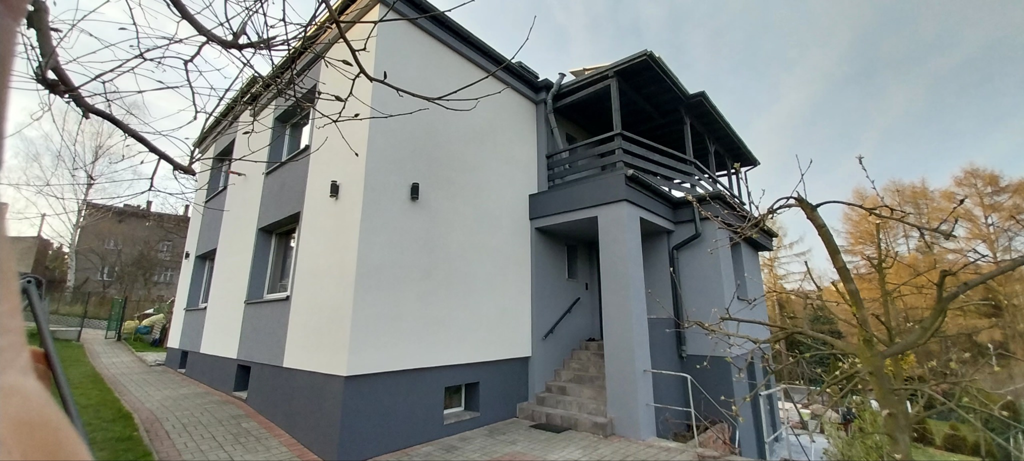 Dwukondygnacyjny dom z balkonem, świeżo po malowaniu elewacji w odcieniach szarości i bieli, z widocznymi detalami architektonicznymi i elementami zieleni w tle.