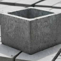 Szary pustak szalunkowy z betonu, kwadratowy z otworem, ułożony na palecie innych pustaków, widoczne logo producenta Wibet.